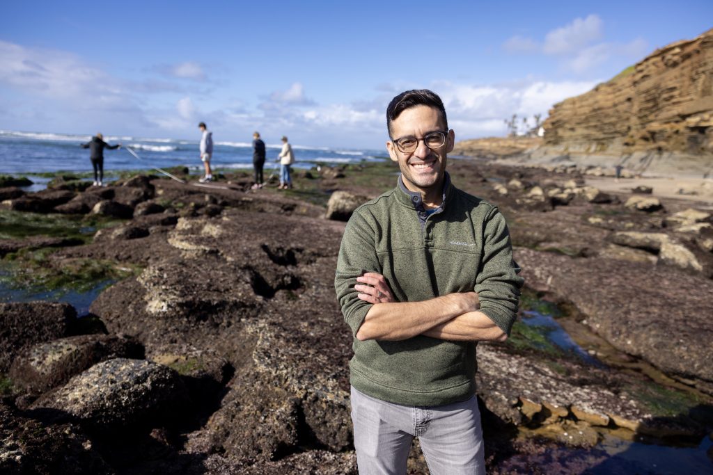 A professor stands with arms folded with tide pools and students exploring them in the background