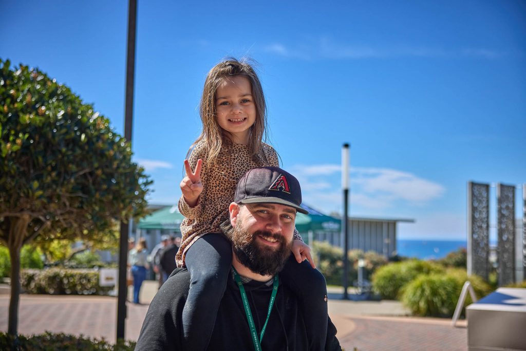A girl sits on her dad's shoulders and gives a smile and a peace sign.