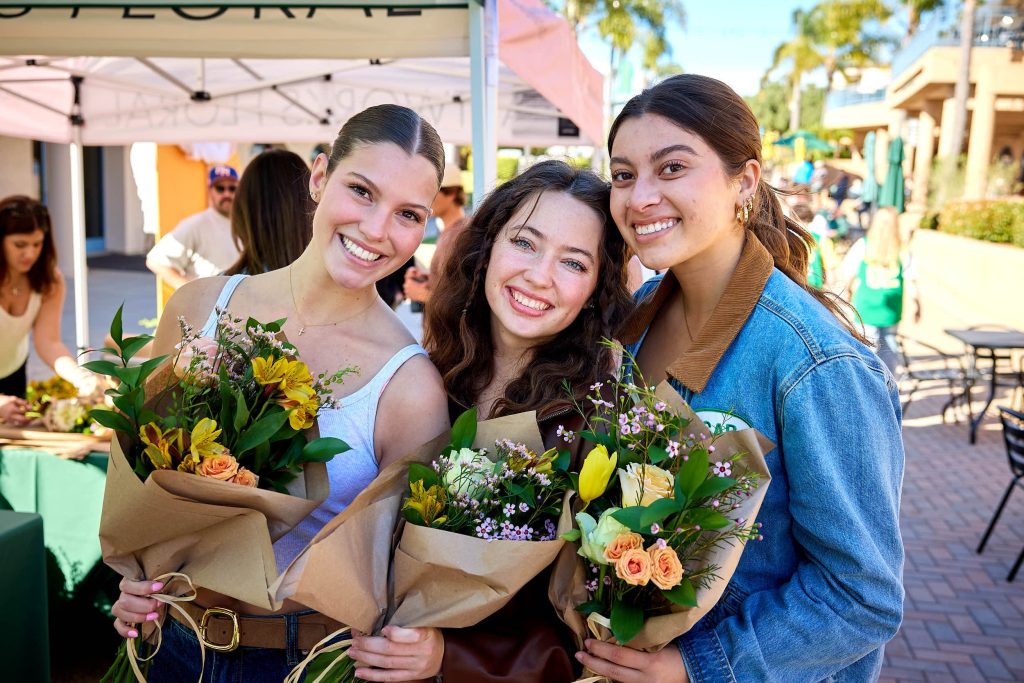 Three PLNU women students gather close for a photo while holding bouquets of flowers.