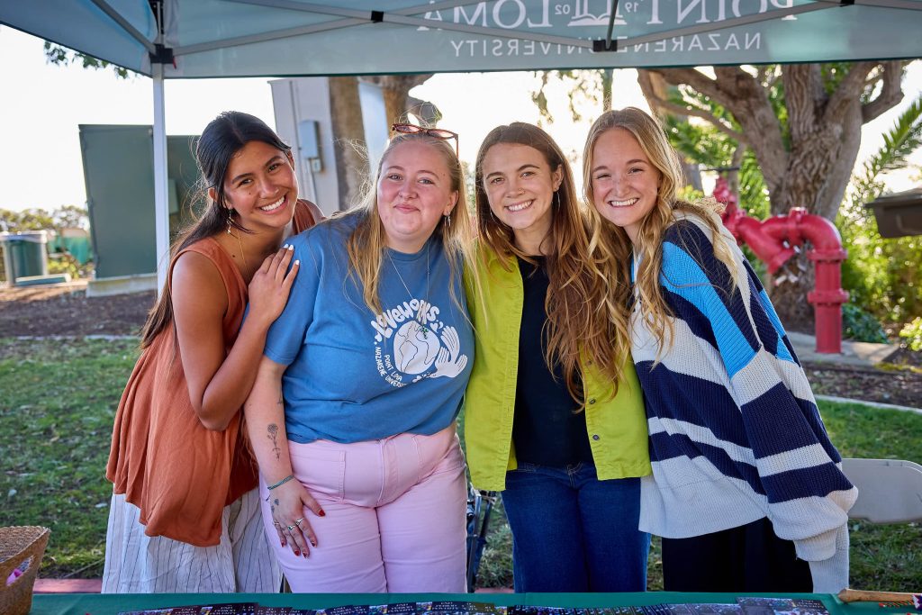 A group of PLNU female students stand behind a booth at PLNU Homecoming.