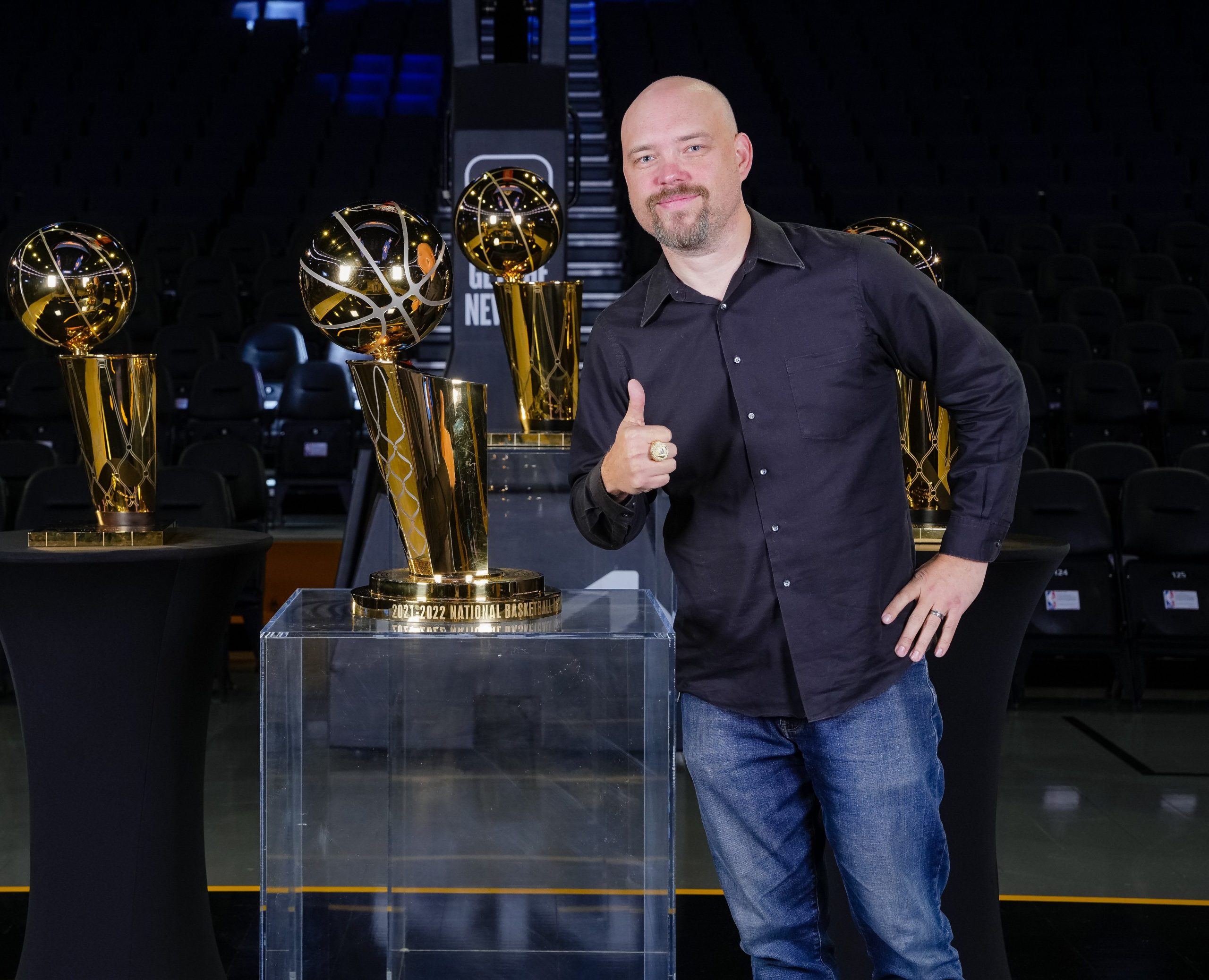 Heitman standing with a thumbs up in front of a few of the championship trophies. 