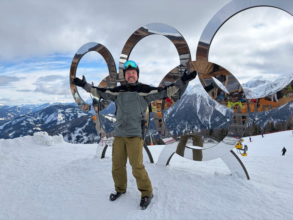Heitman stands with his arms open wide in the snow in front of the silver mirrored Olympic rings. 