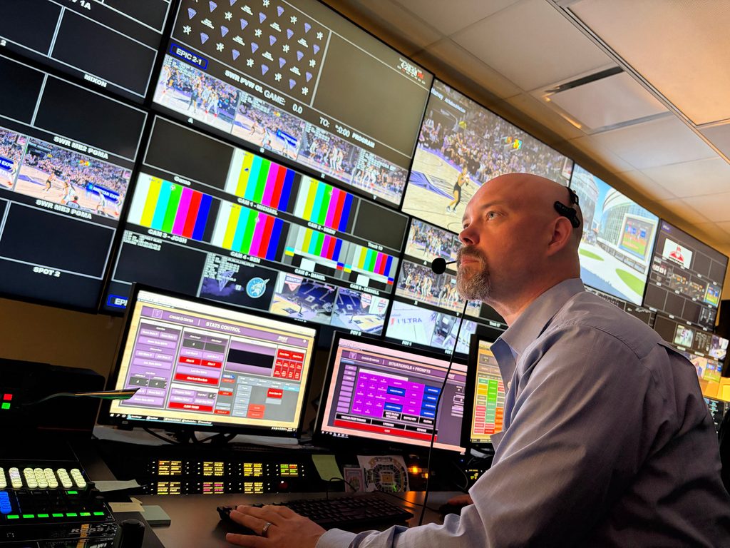 Heitman sitting with a headset in front of a big wall of screens, computers, and other broadcasting equipment. 